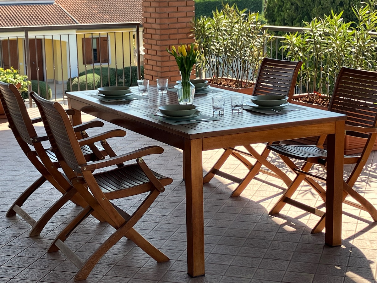 A wooden outdoor dining table is set with plates and glasses, surrounded by four folding chairs. Lush greenery and a railing provide a natural backdrop, while sunlight filters through the area, creating a welcoming space for meals.