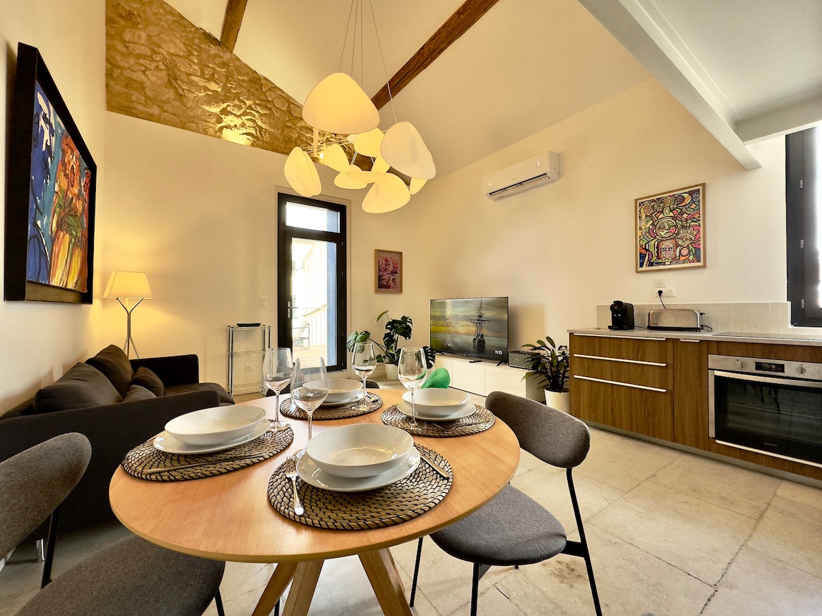 A modern living area features a wooden dining table set for four, surrounded by stylish chairs. A well-equipped kitchen appears in the background, complemented by a flat-screen TV and artistic decor. Natural light fills the space, highlighting the high ceiling and exposed beams.
