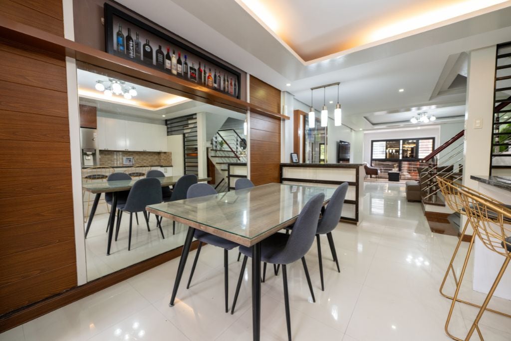 A modern dining area features a stone-topped table surrounded by upholstered chairs. Bright lighting illuminates the space, which includes a mirrored wall and a stylish bar area. A staircase can be seen in the background, adding depth to the open layout.