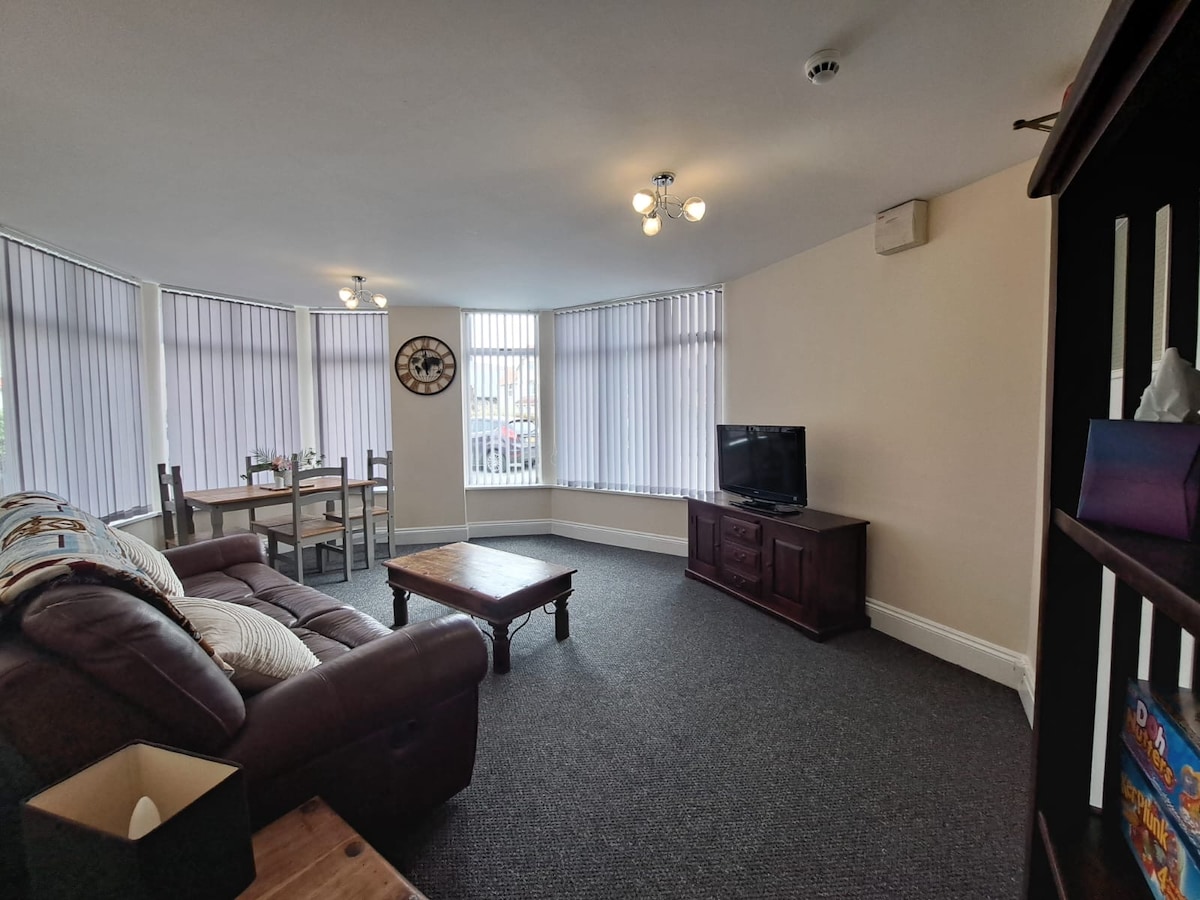 The spacious lounge area features a comfortable sofa, a coffee table, and a TV set. Natural light enters through large windows adorned with vertical blinds. A dining table for four is situated in the corner, enhancing the inviting ambiance of the room.