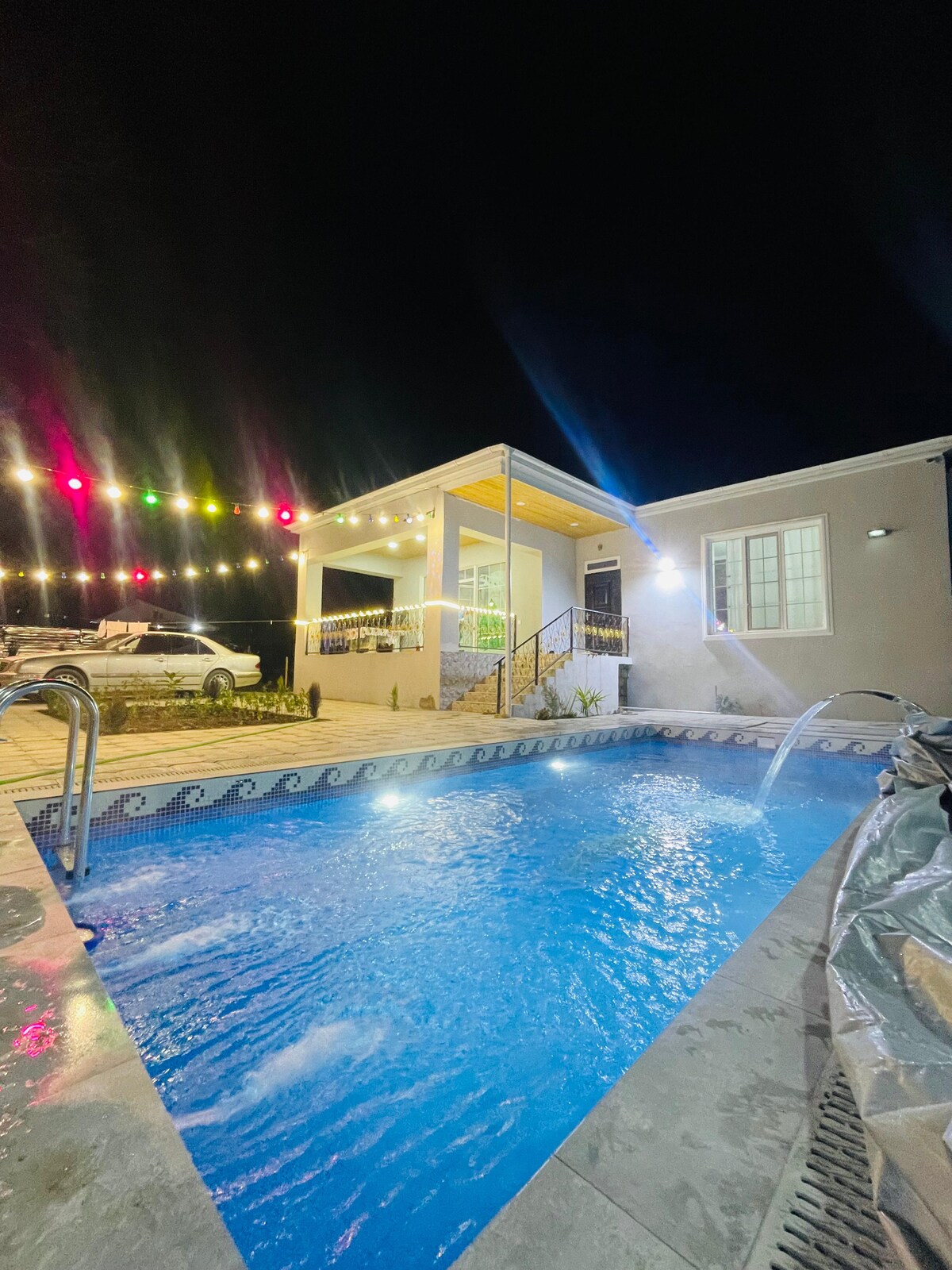A modern home is illuminated at night, featuring a sparkling blue pool with water features. Soft lighting from decorative string lights enhances the outdoor area. A cozy porch is visible, and a vehicle is parked nearby, adding to the welcoming setting.