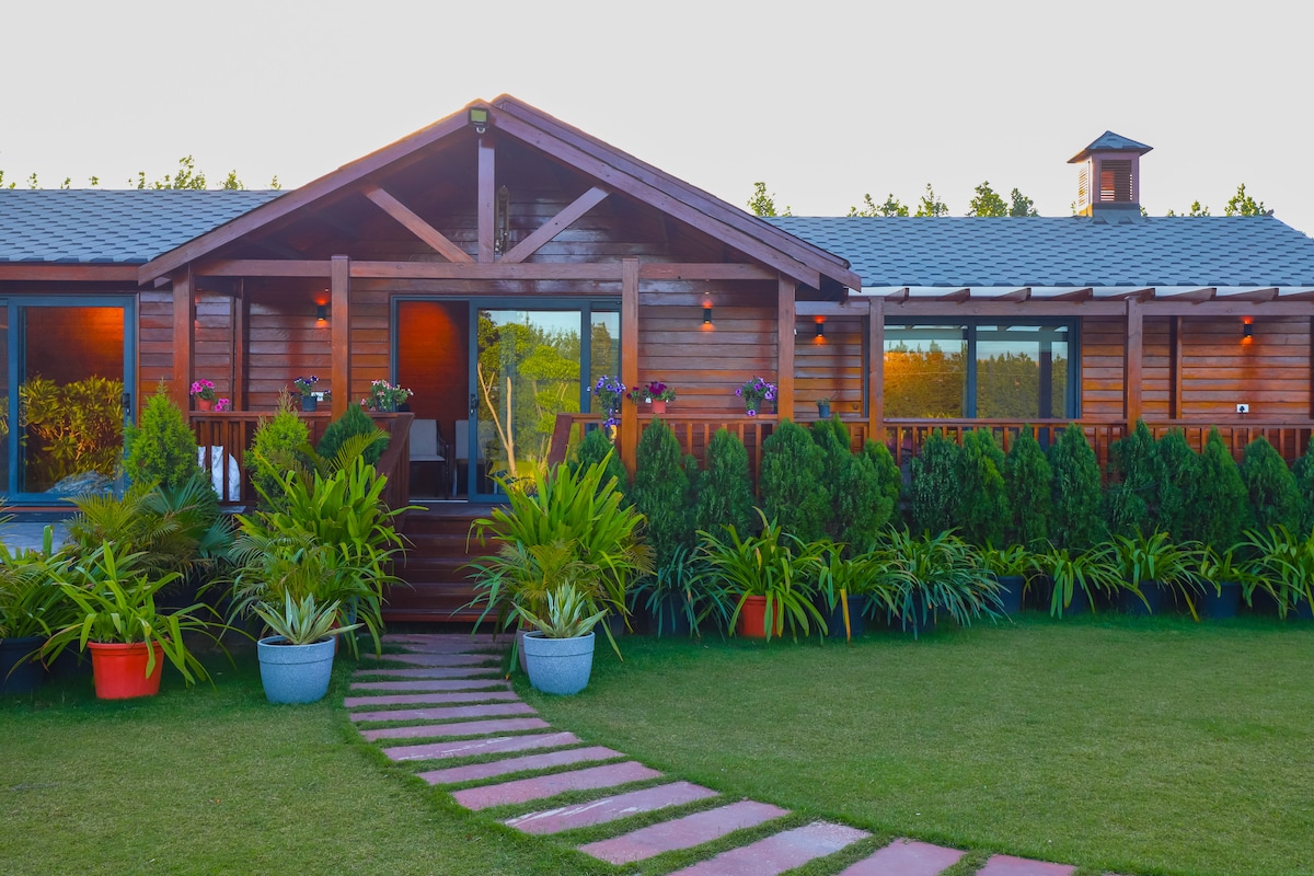 A wooden facade defines the entrance of the farmhouse, featuring large glass doors and a charming porch. Lush greenery surrounds the property, with potted plants lining the pathway leading to the entrance, emphasizing the vibrant garden setting.