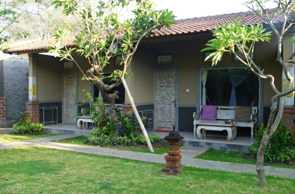 A charming outdoor area features well-maintained greenery, with flowering plants and neatly arranged pathways. Two separate entrances are visible, each flanked by wooden benches. Natural light highlights the textured walls and rustic doors, set against a roof with traditional tiles.