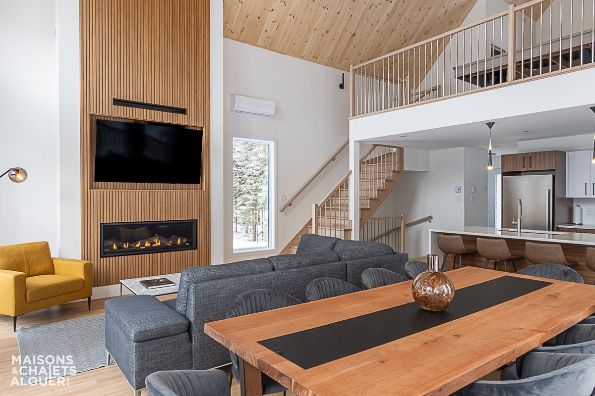 An open living area features a modern design with a comfortable seating arrangement facing a wall-mounted TV above a sleek fireplace. A large wooden dining table is set for gatherings, while a bright yellow armchair adds a pop of color. The wooden ceiling creates a warm ambiance.