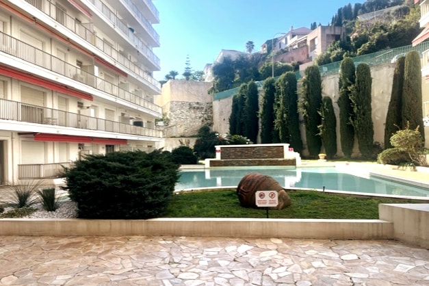 The inner courtyard features a tranquil water basin surrounded by landscaped greenery and tall coniferous trees. A stone pathway leads to the water, with a grassy area and decorative signage present. The backdrop includes a multi-story building, contributing to a peaceful ambiance.