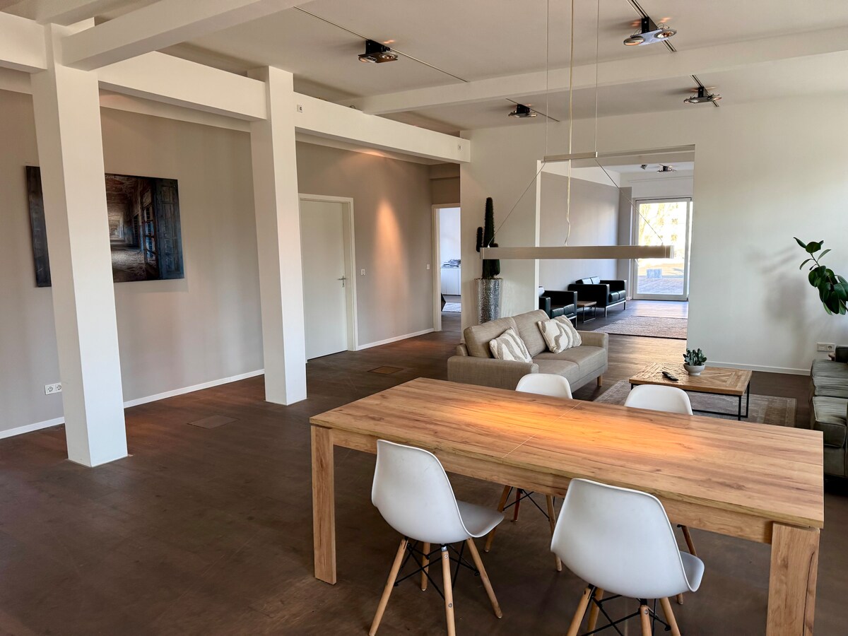 An open living space features a wooden dining table with white chairs, set against a backdrop of light-toned walls. A comfortable sofa and lounge chairs are arranged in the adjacent area, inviting relaxation. Natural light fills the room through large windows.