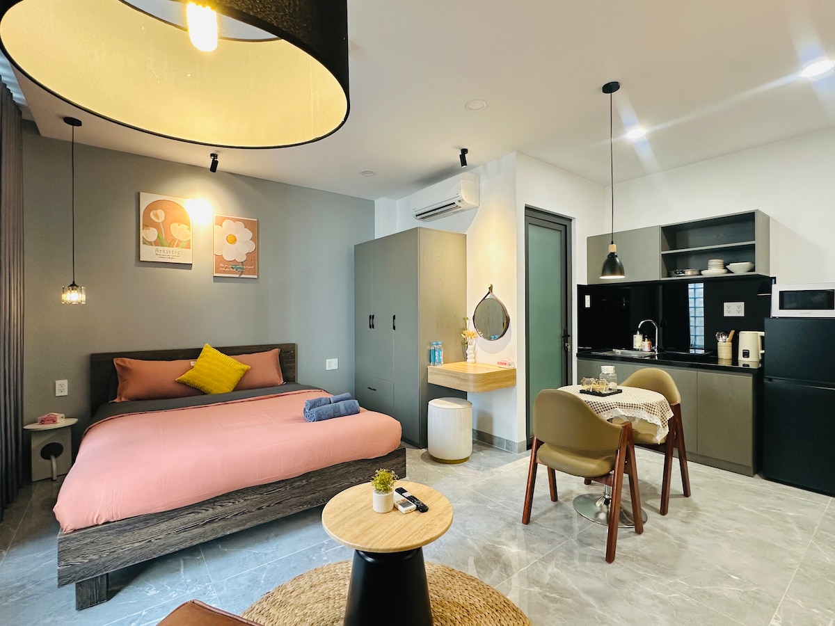 An open-plan living area features a spacious bed dressed in soft pink linens. A round dining table with four chairs is positioned beside a kitchenette, which includes modern appliances. Decorative wall art and overhead pendant lights add warmth to the neutral tones of the space.
