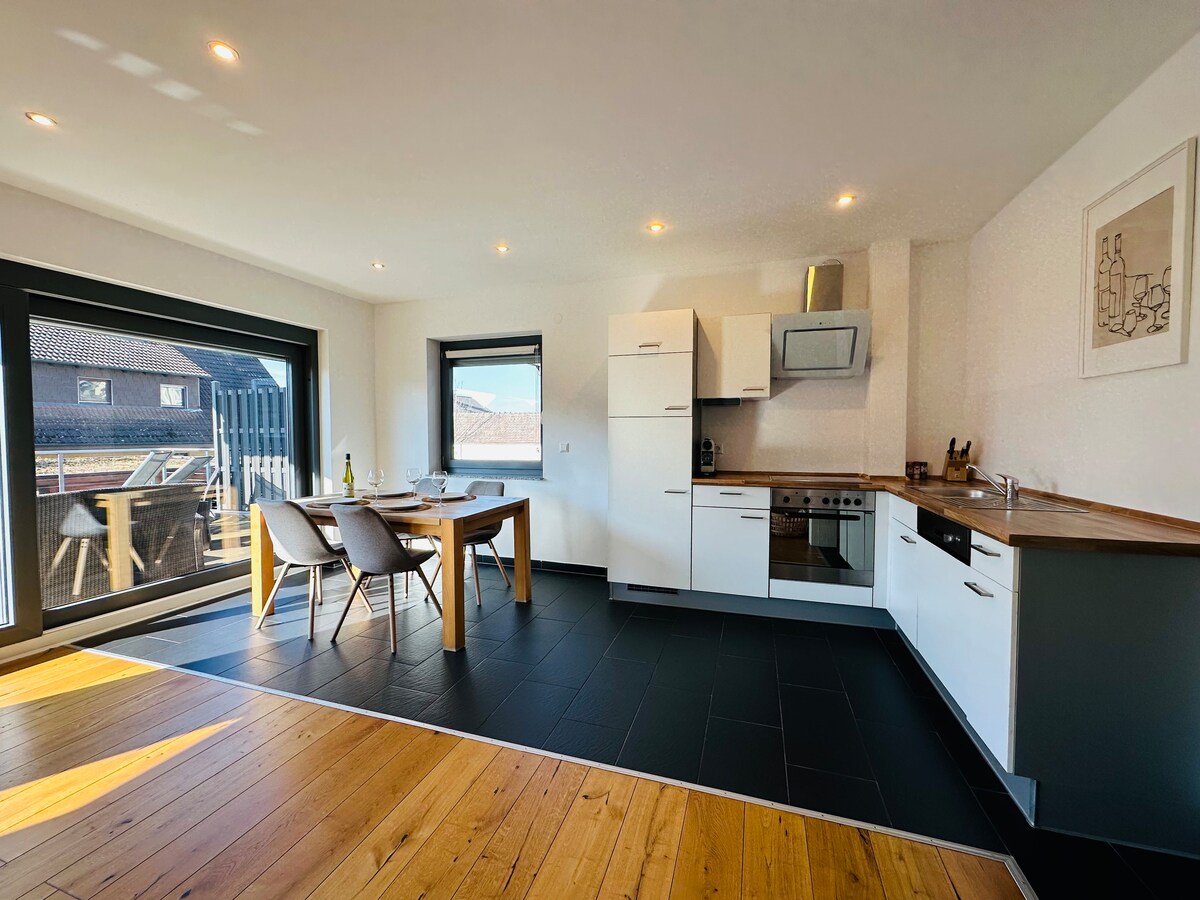 A modern kitchen and dining area features sleek white cabinets and wooden countertops. A dining table is accompanied by four chairs. Large glass doors provide natural light and access to a balcony, while a built-in oven and stovetop complete the well-equipped kitchen.