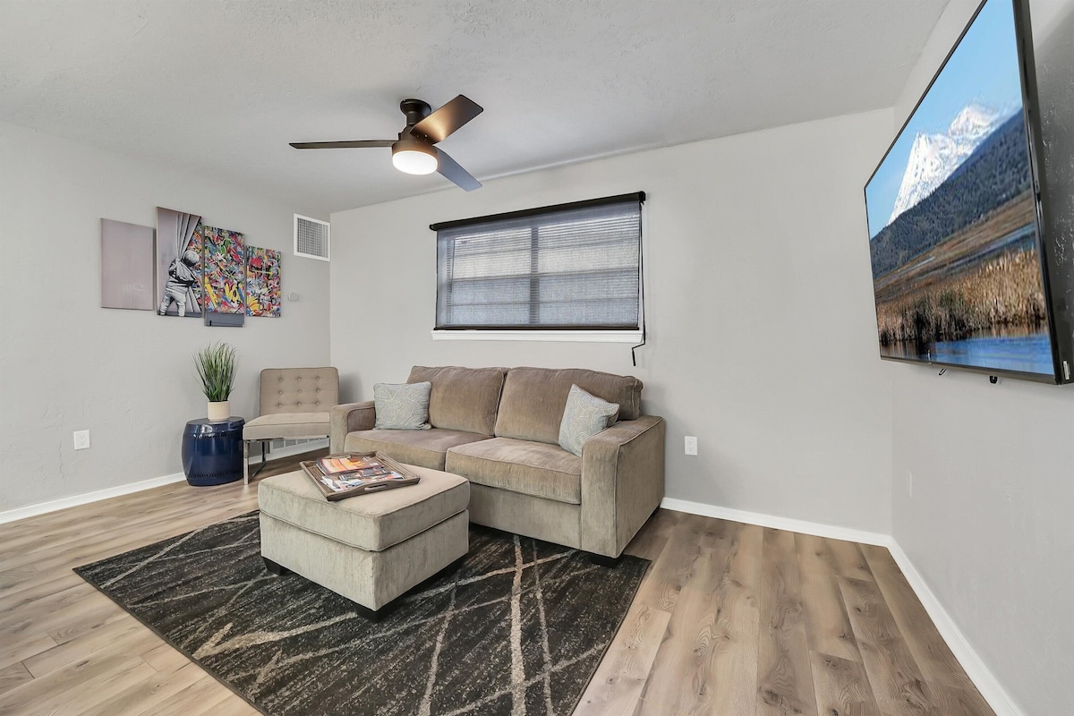 A modern living area is presented with a sectional sofa and an ottoman centered on a textured rug. A flat-screen television is mounted on the wall, while natural light filters through a partially opened window, complemented by neutral wall colors and artistic decorations.