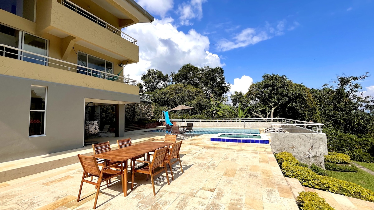 An outdoor dining area is positioned on a stone patio, featuring a wooden table with several chairs. A private pool glistens nearby, bordered by lush greenery and mountain views under a clear blue sky.