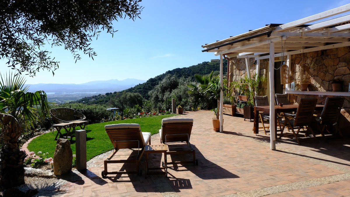 A sunlit patio area is shown, featuring wooden lounge chairs positioned for relaxation. Lush greenery and a landscaped garden fill the background, while distant hills and mountains are visible in the horizon. A rustic dining area under a covered structure offers an inviting space for outdoor meals.