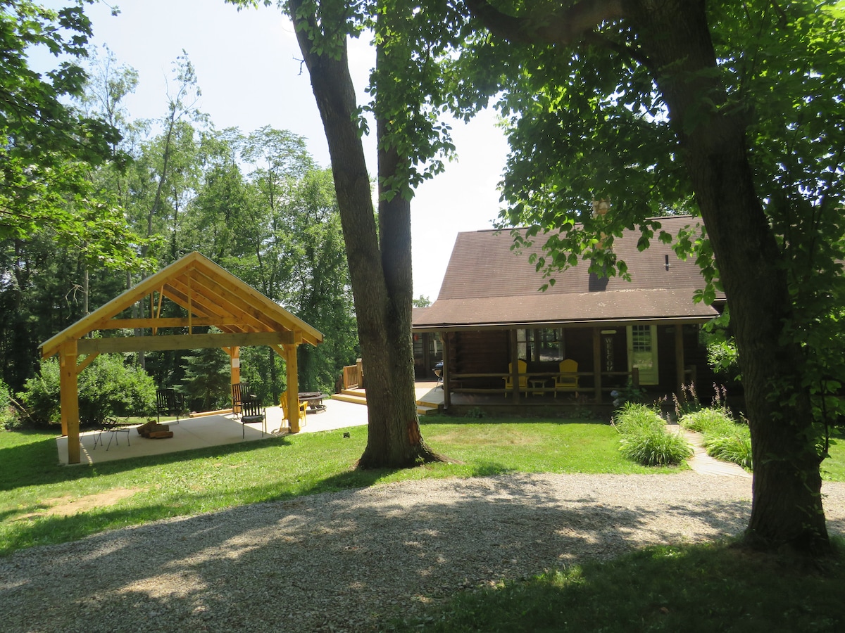 A log cabin is set among lush greenery, featuring a covered outdoor seating area constructed of wood. The cabin's porch is adorned with yellow chairs, inviting relaxation. The surrounding landscape includes tall trees and bushes, providing a serene and natural environment.
