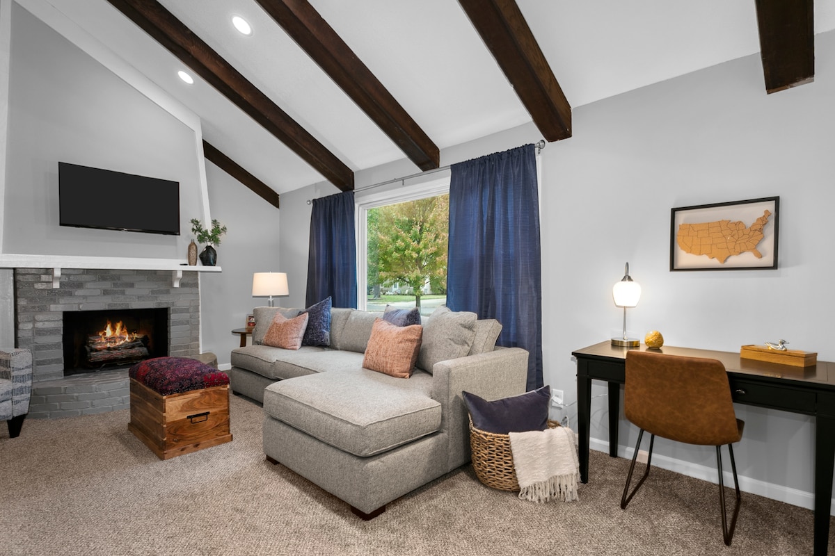 The living room features vaulted ceilings with exposed wooden beams. A sectional sofa is complemented by decorative pillows, and a cozy fireplace is situated nearby. Large windows allow natural light to fill the space, while a small desk with a chair is positioned against the wall.
