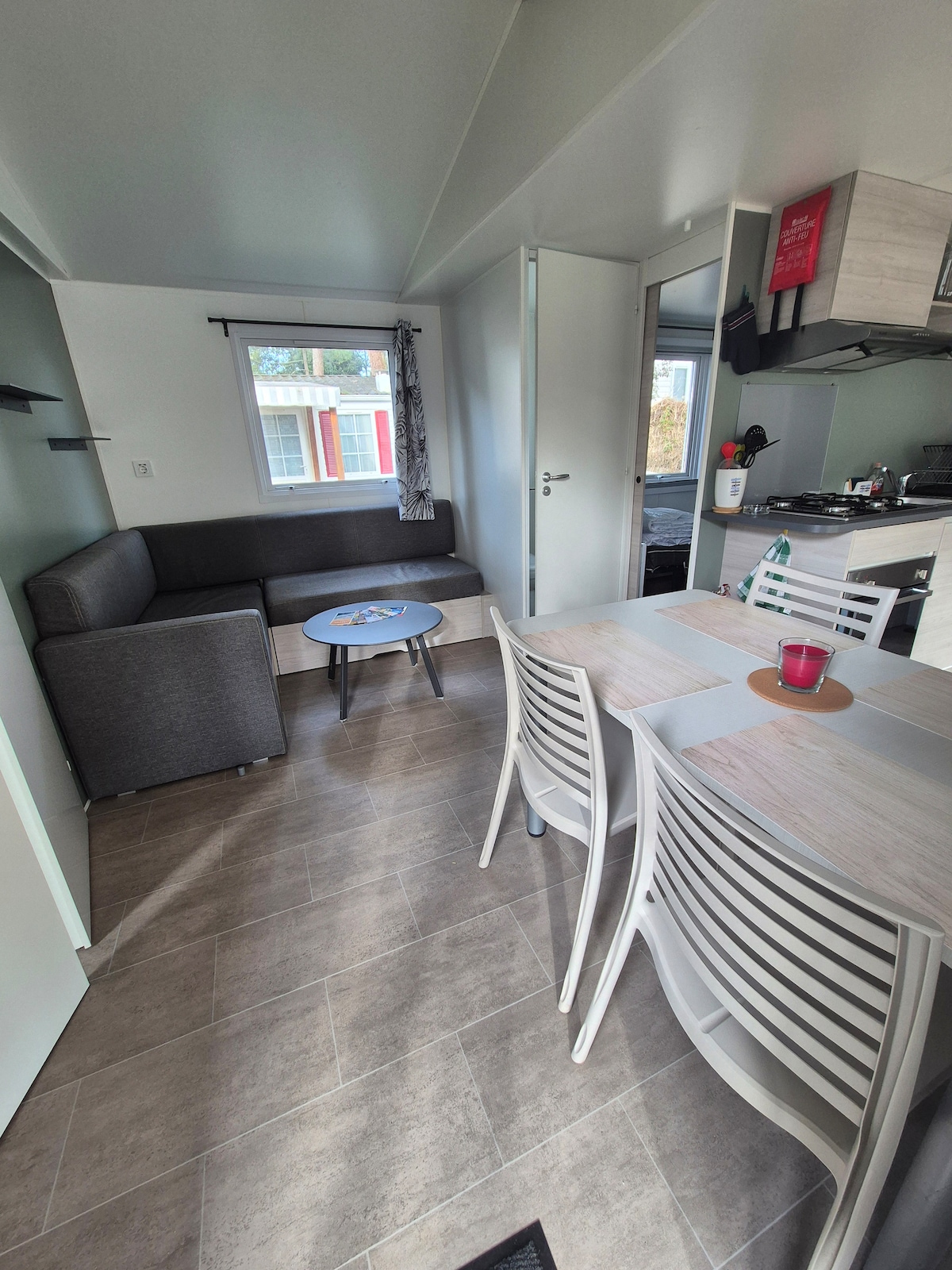 A living area is depicted with a gray sofa positioned near a circular table. Light-colored chairs surround the table. A doorway leads to another room, while a window allows natural light to illuminate the space, providing a view of the outside.