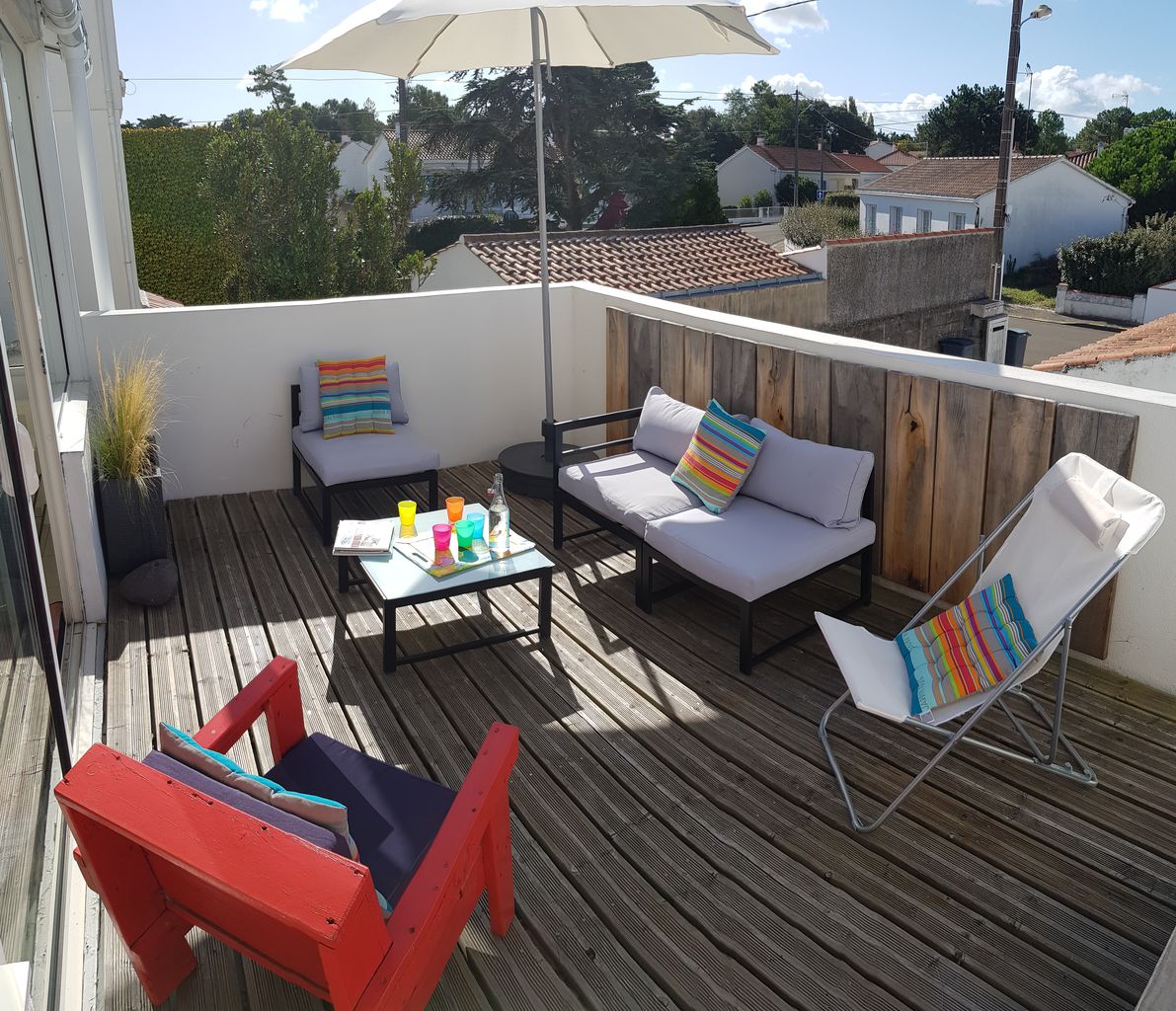 A spacious outdoor terrace is furnished with casual seating, including two lounge chairs and a low table. A large umbrella provides shade, while colored cushions add a vibrant touch. Wooden decking surrounds the area, creating a warm and inviting space for relaxation.