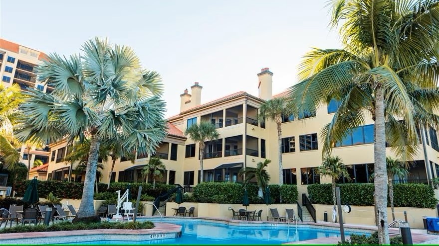 The exterior of the condo complex is visible, surrounded by lush palm trees and tropical landscaping. A swimming pool is positioned in the foreground, with sun loungers placed nearby. The building features multiple windows and balconies that offer a relaxed vacation atmosphere.