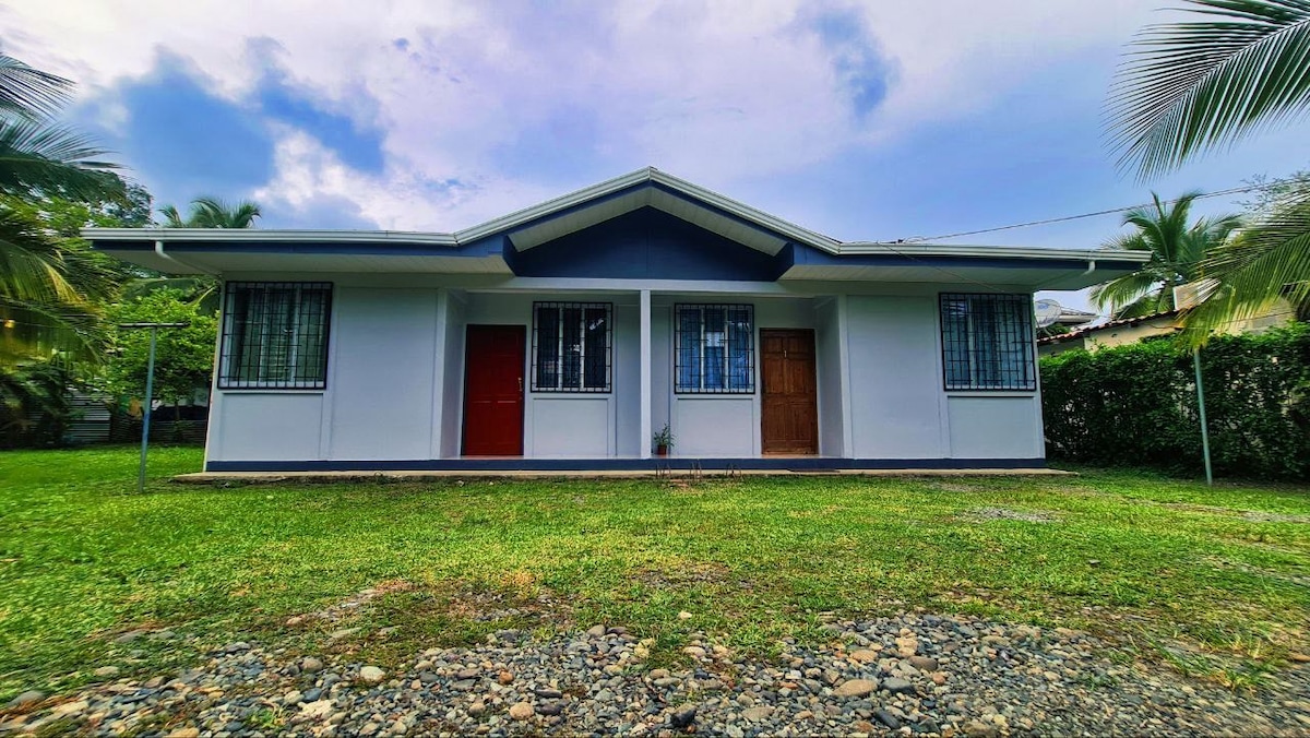 A single-story structure is visually divided into two units, featuring large windows with metal grilles. The building is painted in light colors, complemented by a contrasting dark roof. A well-kept grassy area surrounds the front, with palm trees framing the view.