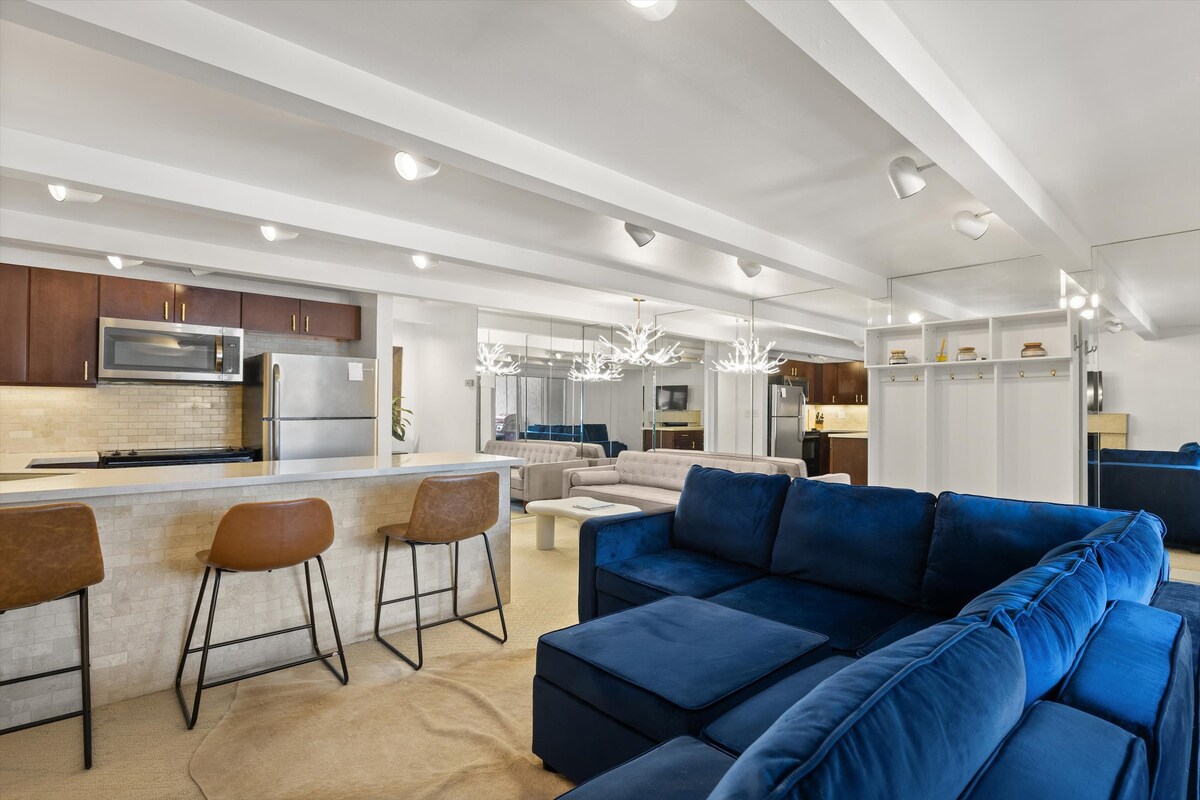 An open living area features a spacious blue sectional sofa with an ottoman, arranged in front of a kitchen bar with three modern stools. Soft lighting from stylish fixtures illuminates the area, while mirrored walls enhance the sense of space.