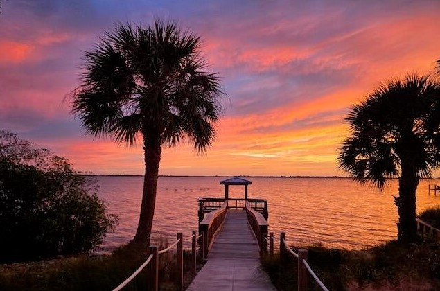 A wooden dock leads towards the water, framed by palm trees. The sky is filled with vibrant hues of orange, pink, and purple as the sun sets over the tranquil surface of the river.