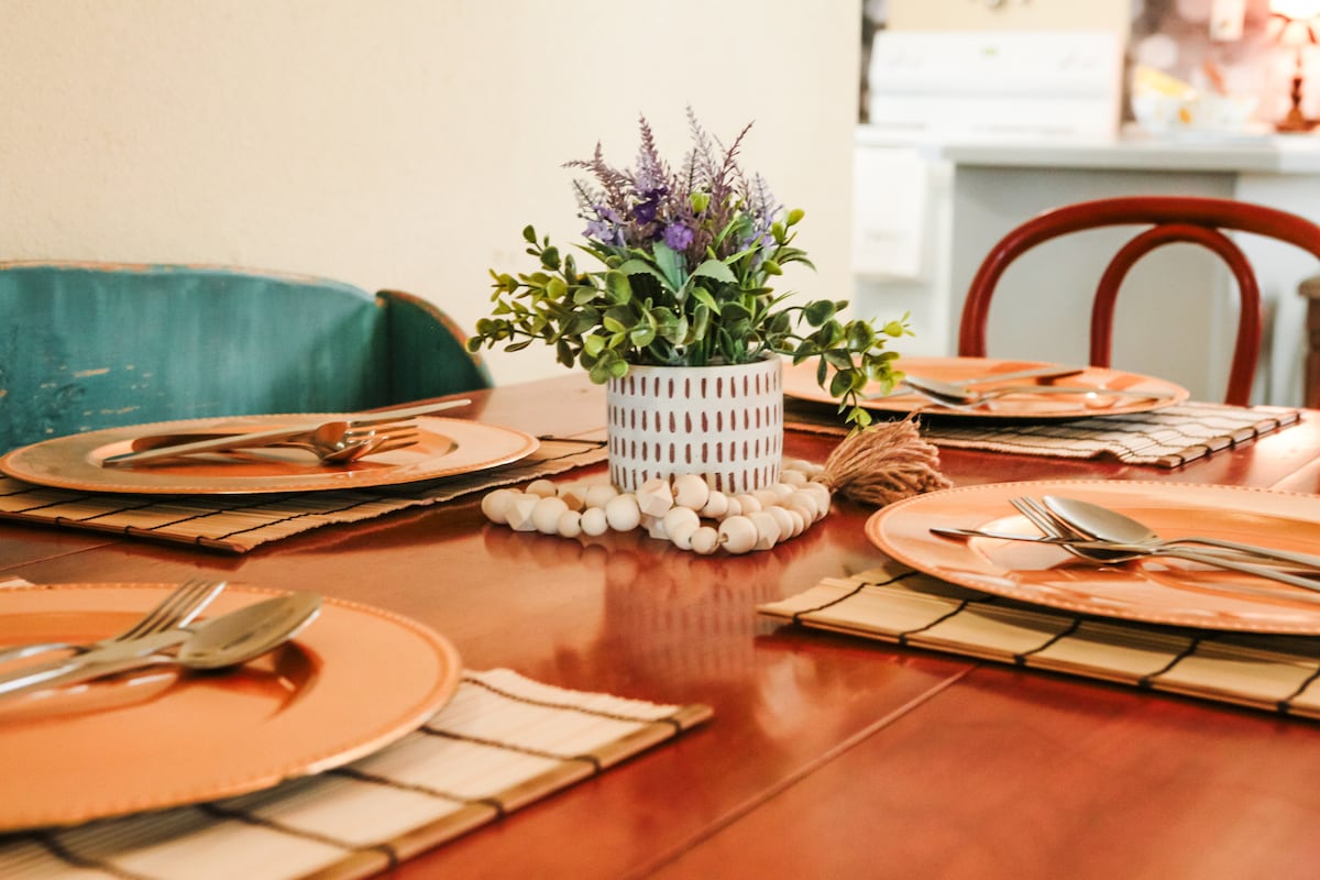 A dining table is set with elegant plates and utensils for four, showcasing metallic tones. A decorative arrangement of greenery and small white accents sits at the center. The background indicates a glimpse of the kitchen area with soft lighting.