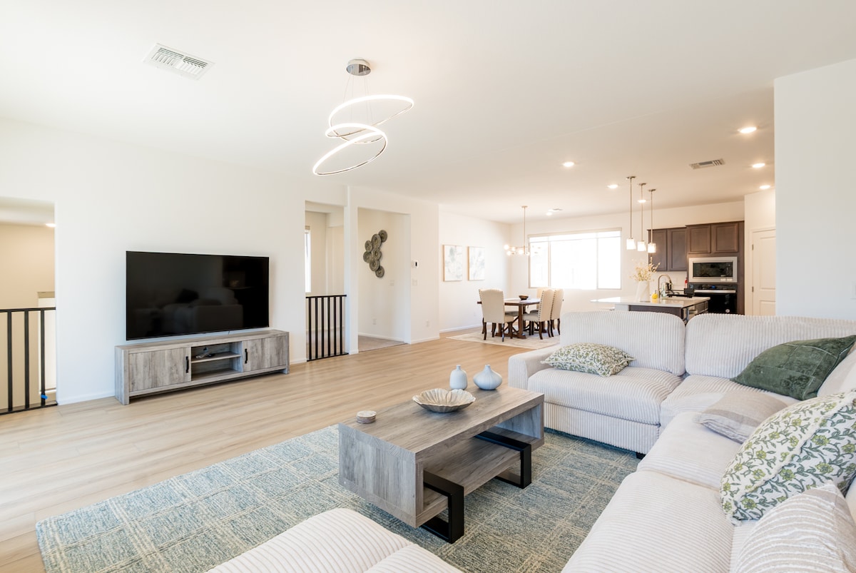 A spacious living area is highlighted by a large sectional sofa adorned with decorative pillows. A flat-screen TV is mounted on a light wood entertainment center. Natural light fills the room, creating a bright ambiance. A dining area and modern kitchen are visible in the background.