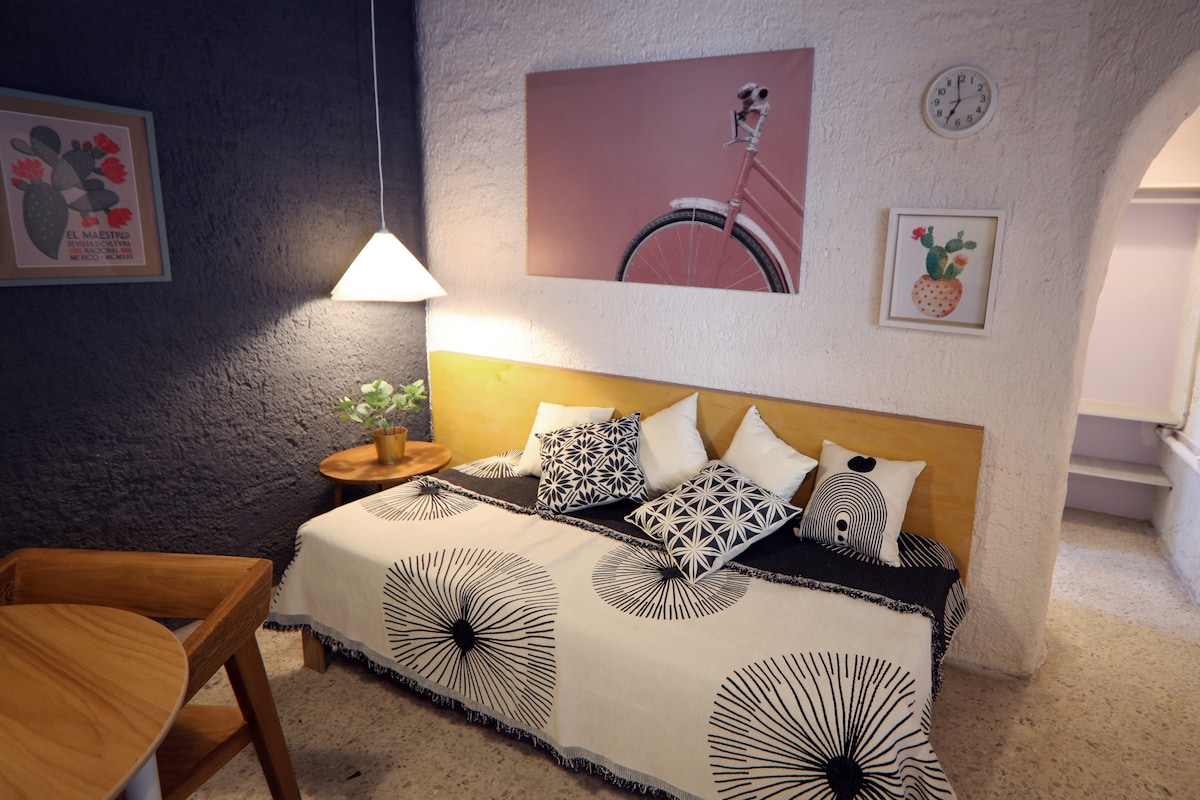 A compact living space is defined by a wooden bed adorned with a black and white patterned blanket and pillows, accompanied by a small round table. Bright wall art and a clock are displayed above, while a gentle light is emitted from a standing lamp nearby.