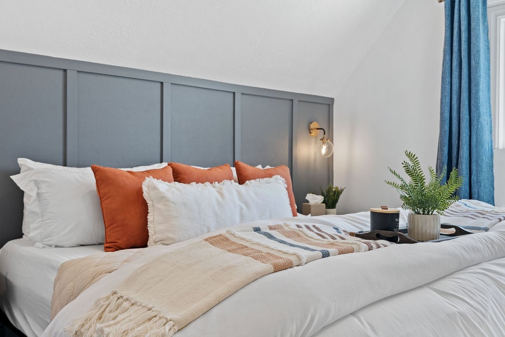 The bedroom features a medium-firm king-sized bed with white linens accented by orange and neutral throw pillows. A cozy blanket is draped across the bed, and a small tray holds decorative items and a potted plant. Soft lighting and curtains enhance the serene atmosphere.