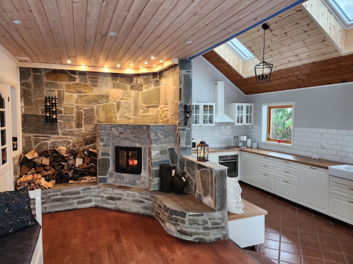 An inviting living area features a prominent stone fireplace, with firewood neatly stacked beside it. Natural light floods the space through skylights and large windows. A cozy seating nook with cushions is positioned under the window, complemented by a well-equipped kitchen visible in the background.
