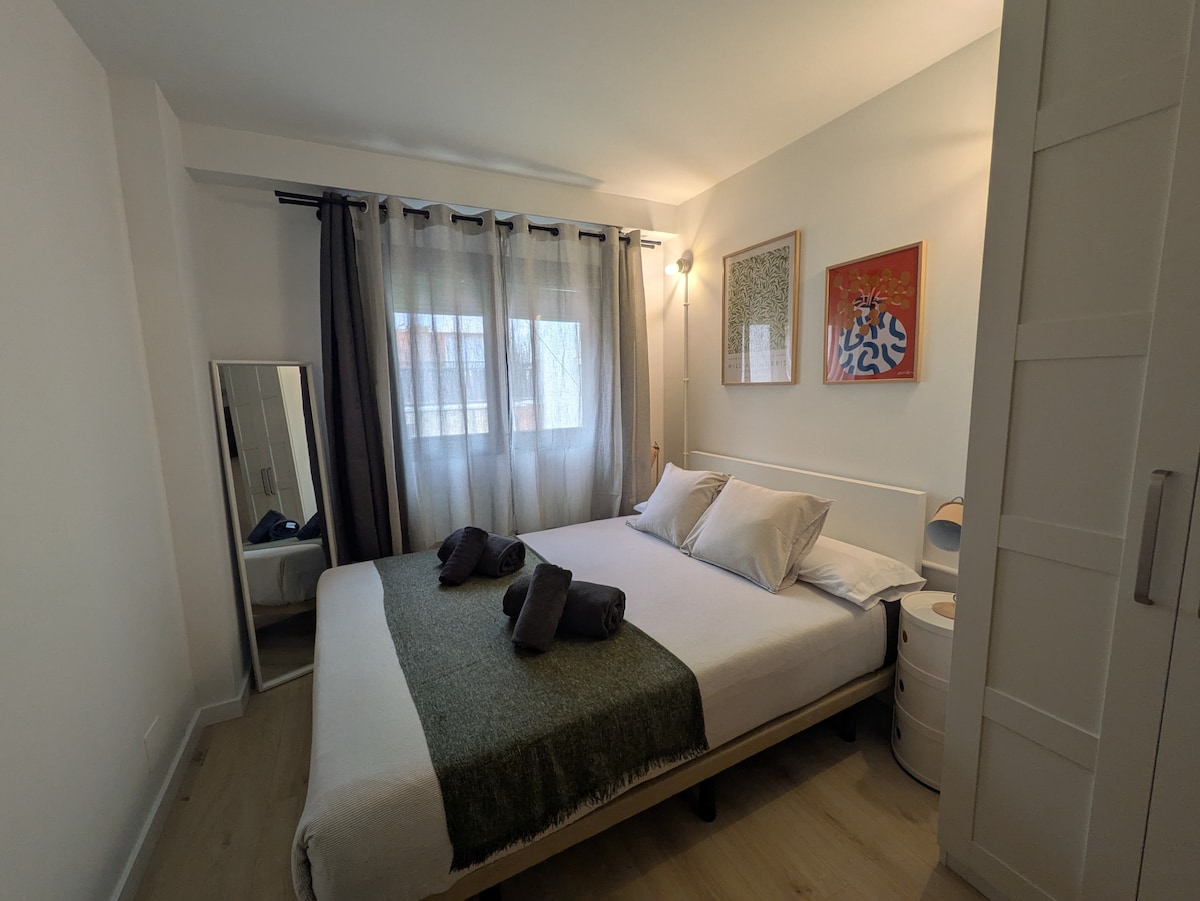 A spacious sleeping area features a cozy double bed with a light-colored duvet and decorative pillows. Soft light enters through a partially covered window, while a mirror and a nightstand are visible. The room's design is complemented by modern wall art.