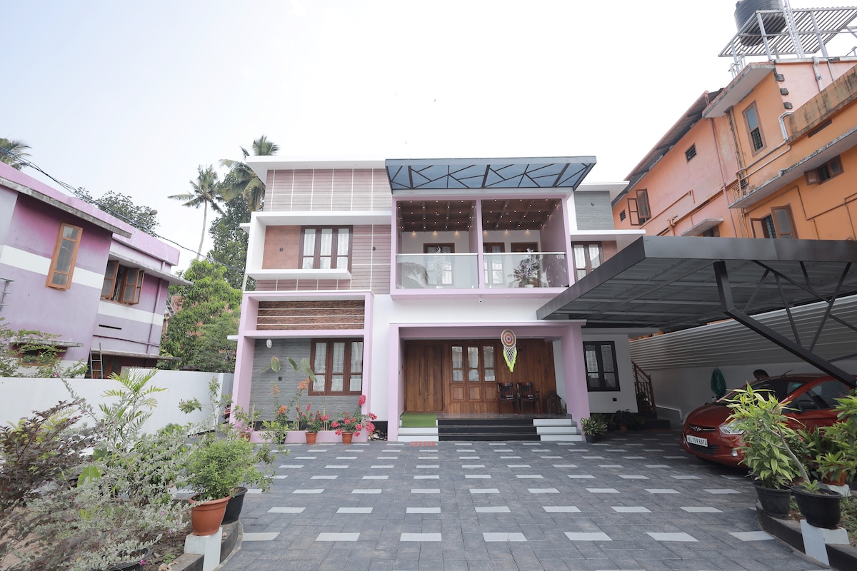 The exterior of a modern two-story building is shown, featuring a mix of colors with a prominent pink hue. A spacious driveway is visible, lined with potted plants. Large windows allow for ample natural light, while a covered parking area accommodates multiple vehicles.