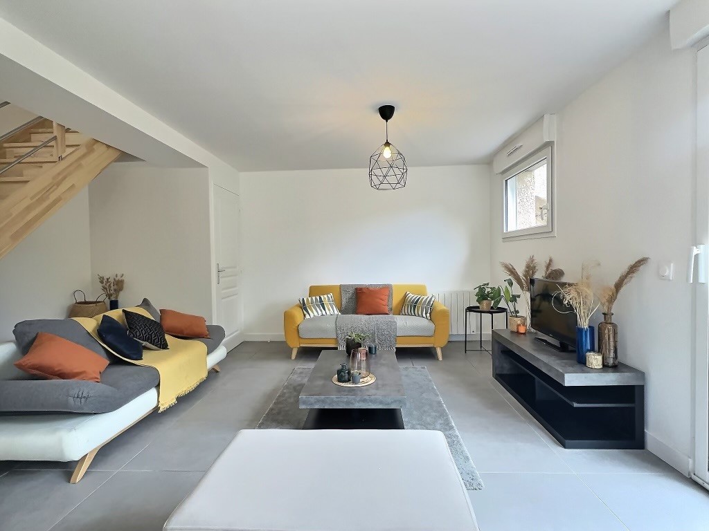 A bright living space is shown, featuring two sofas in contrasting colors, accented with decorative pillows. A low coffee table is placed centrally on a textured rug, surrounded by potted plants. A modern light fixture hangs above, and natural light filters through the window.
