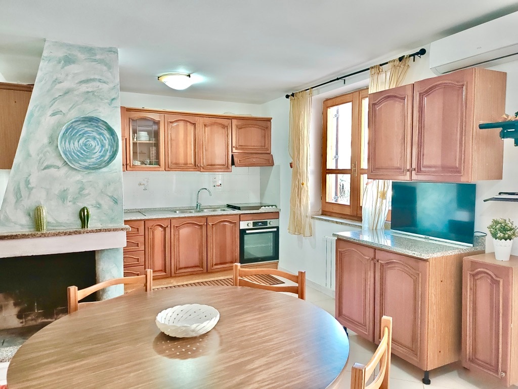 The kitchen area features wooden cabinetry with a built-in oven and a stove, complemented by a refrigerator. A round dining table is set at the center, surrounded by wooden chairs. Natural light enters through a window with sheer curtains, enhancing the inviting atmosphere.
