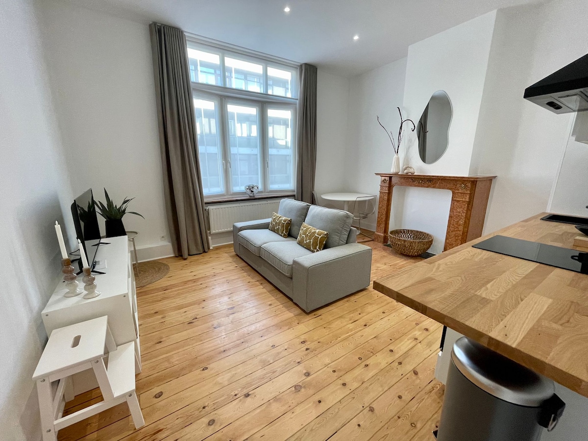 A modern living space features a light grey sofa with decorative cushions and a wooden coffee table. A wall-mounted TV is positioned on a white cabinet. Natural light fills the room through large windows, complemented by neutral curtains and warm wooden flooring.