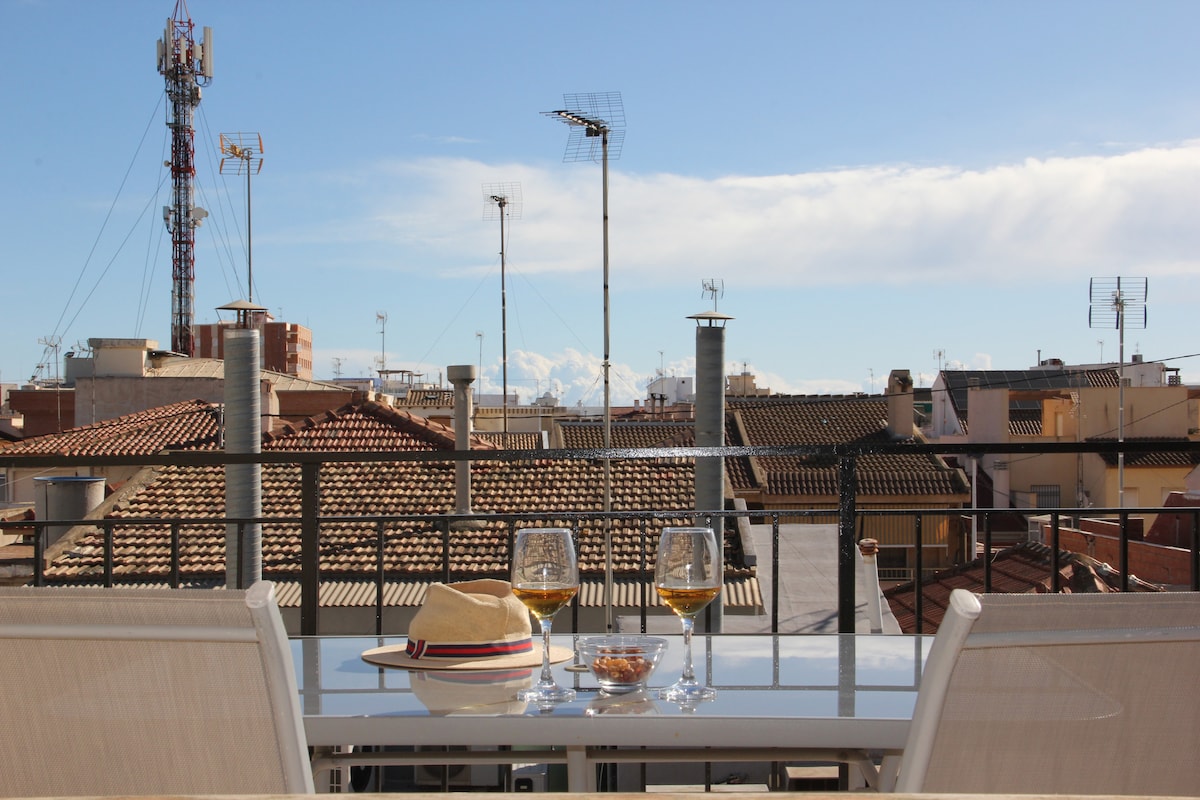 A rooftop terrace is presented with a dining table set for two, featuring glasses of wine and a bowl of snacks. The urban landscape reveals a view of terracotta rooftops and surrounding antennas, with distant mountains visible under a clear blue sky.