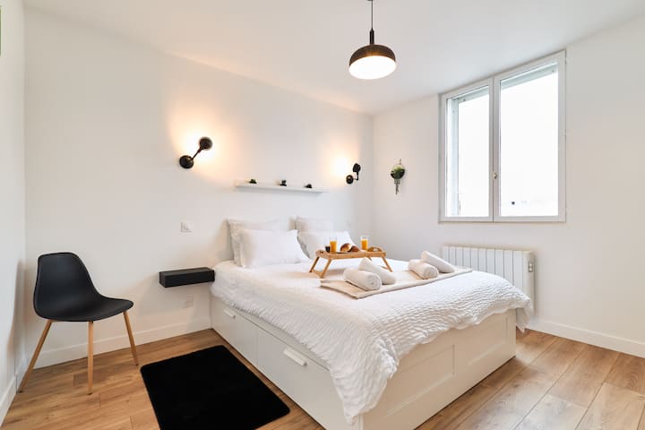 غرفة نوم مشرقة ومرتبة بسرير مزدوج وبياضات بيضاء وتخزين مدمج. الديكور البسيط مع الرفوف والنباتات وتركيبات الإضاءة السوداء يجلب لمسة عصرية.