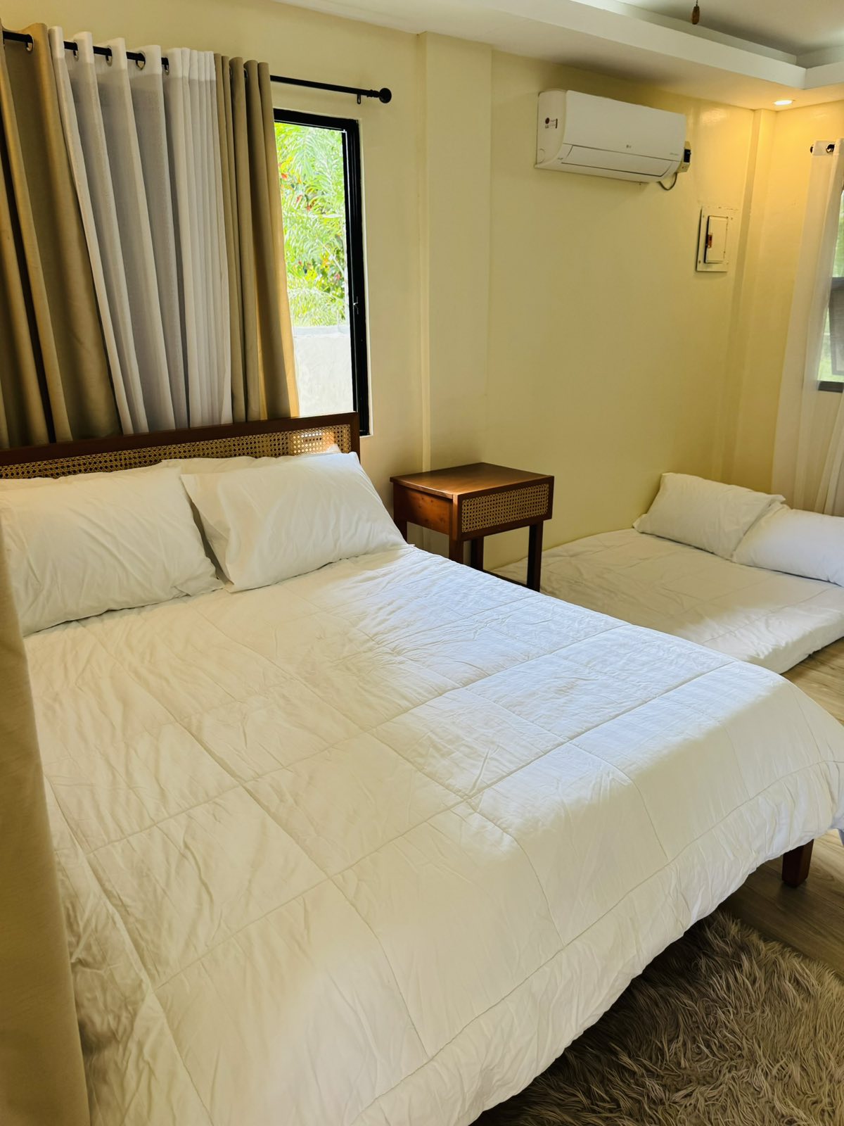 A bright sleeping area features a spacious bed with white bedding, complemented by a cozy single mattress nearby. Natural light floods the room through the window, framed by soft curtains. An air conditioning unit is visible above for climate control.