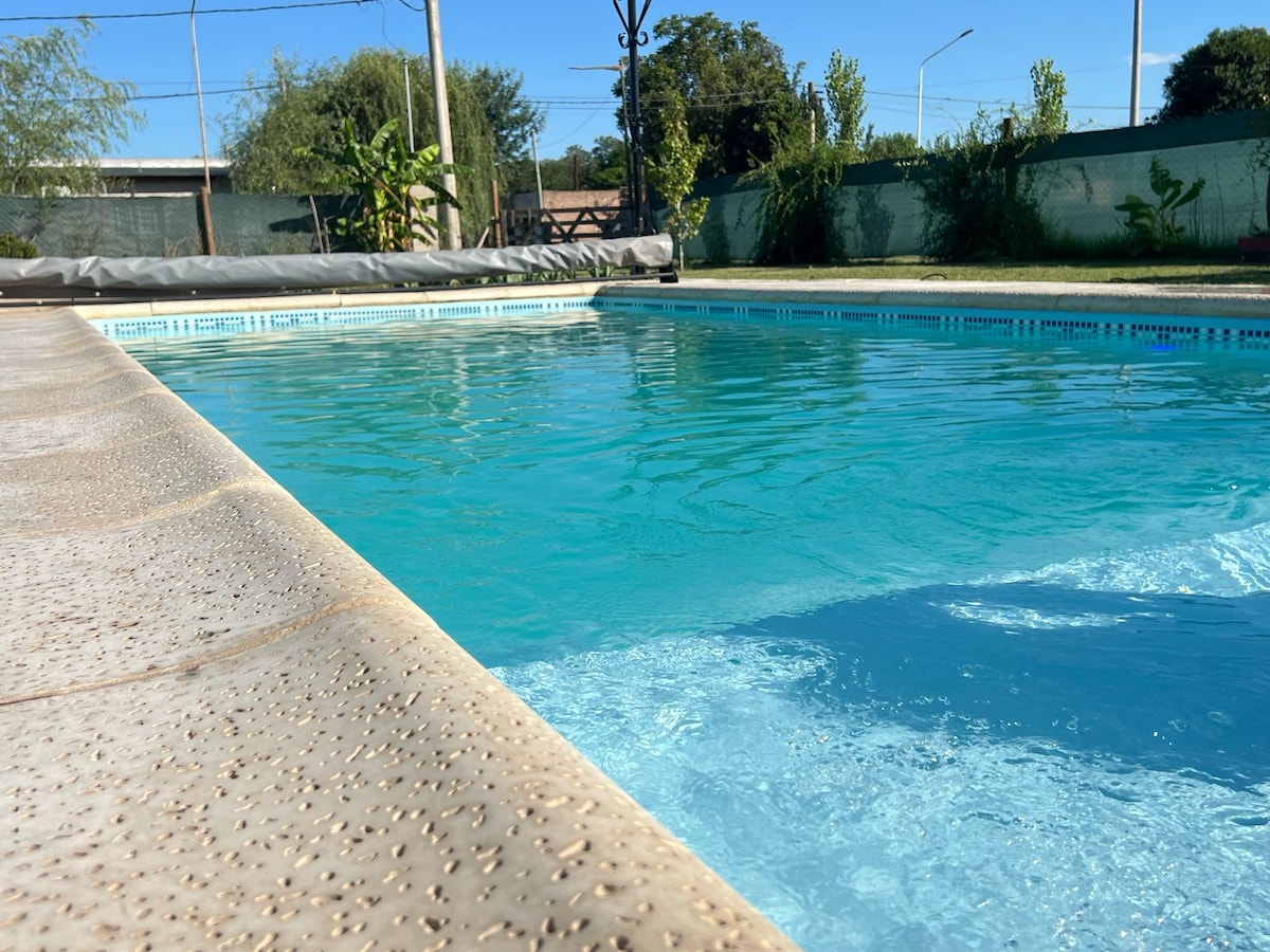 A swimming pool with clear turquoise water is featured, surrounded by a spacious deck. Sunlight reflects off the water's surface, creating a serene atmosphere. Greenery is visible in the background, enhancing the outdoor setting.