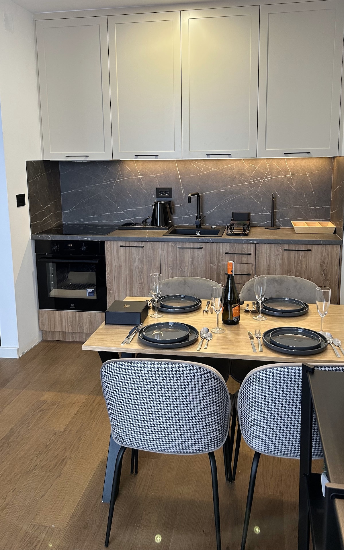 The modern kitchen features sleek cabinetry and a marble backsplash, complemented by stainless steel appliances. A dining table is set for four, with elegant plates and glassware arranged on a wooden surface, inviting guests to gather and enjoy meals together.