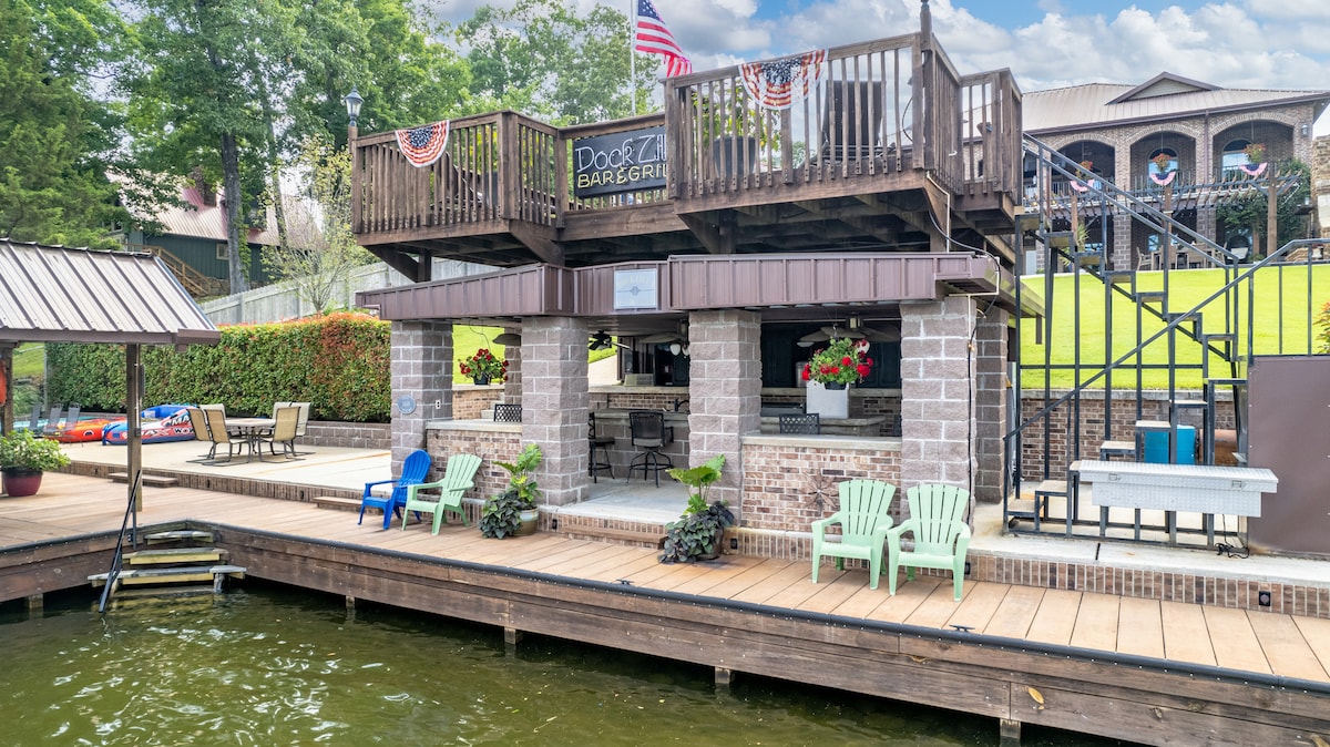 A lakeside outdoor kitchen area features a stone counter with bar stools, surrounded by colorful potted plants. Adirondack chairs are positioned on the wooden deck, providing seating options. A spacious upper deck is visible, adorned with festive decorations and flags.