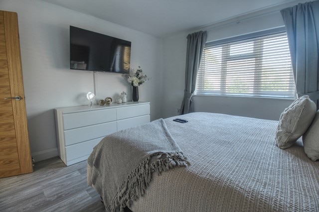 A light-filled sleeping area features a spacious bed adorned with a textured throw. A white dresser stands against the wall beneath a flat-screen television. Window coverings provide adjustable light, while a decorative arrangement on the dresser adds a subtle touch of style.