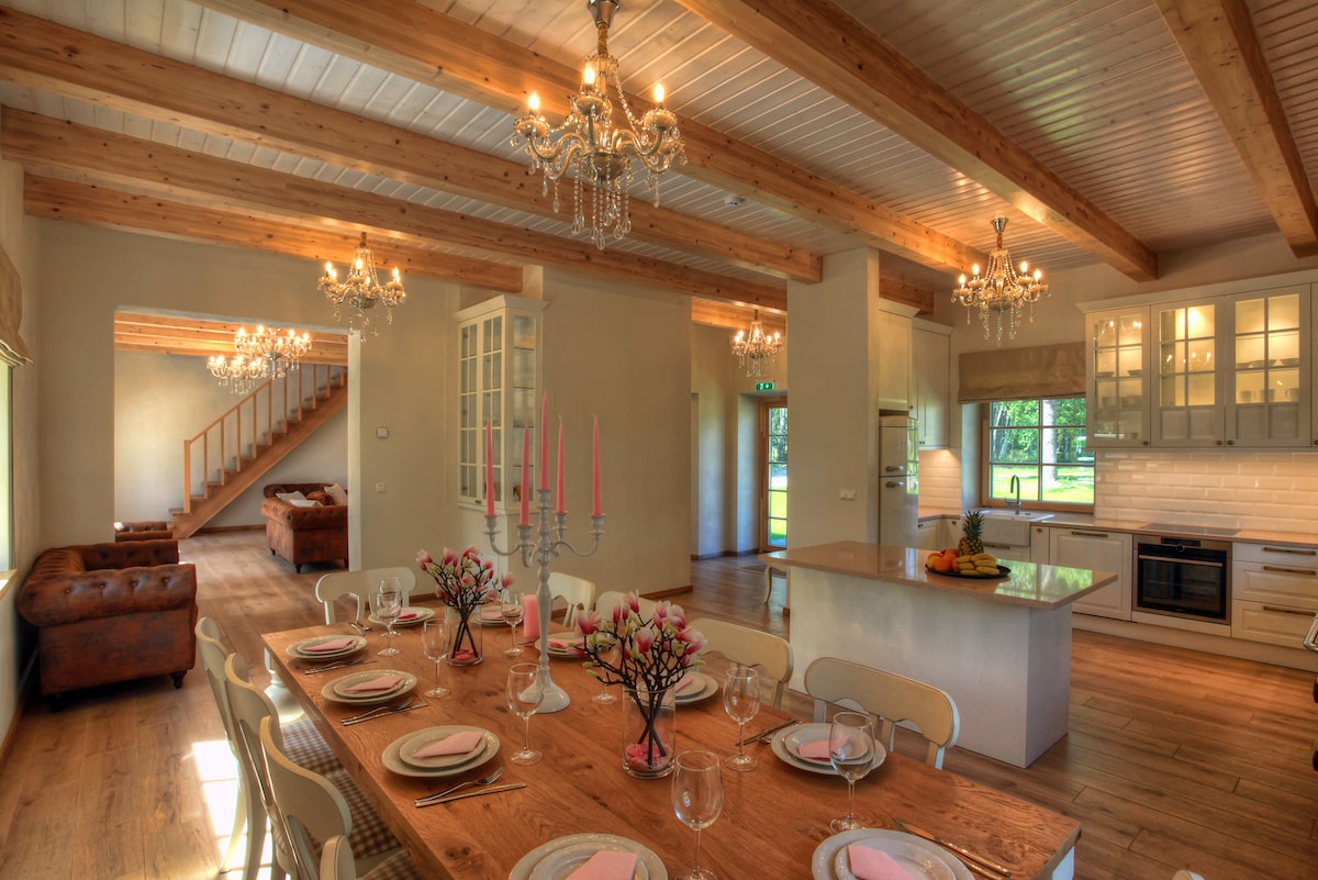 A dining area features a long wooden table set for a meal, accompanied by light-colored chairs. Chandeliers hang from the ceiling, adding elegance. The open layout includes a view of the kitchen and a cozy seating area with a sofa in the background.