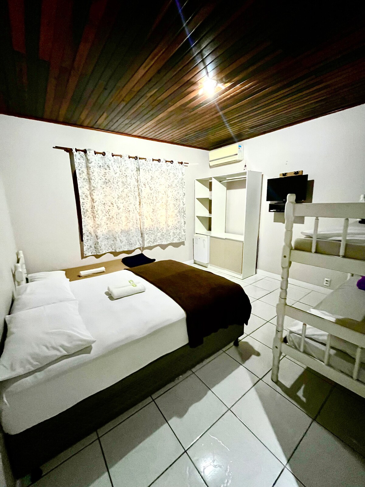 A well-organized guestroom features a spacious bed with white linens and a dark throw blanket. A set of white bunk beds is positioned beside the bed. A window with light-filtering curtains allows natural light to fill the space, complemented by wood-paneled ceilings.