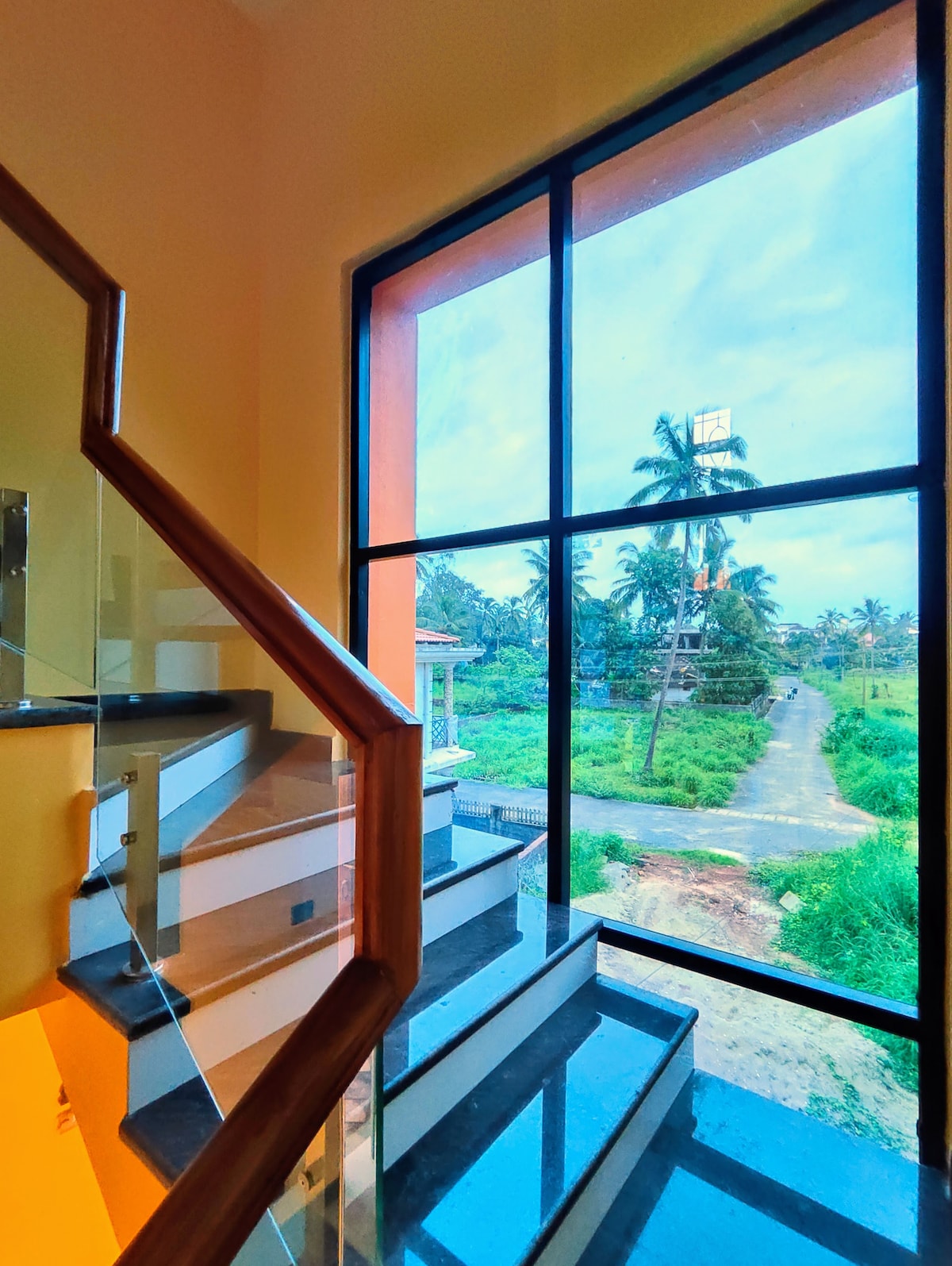 A modern staircase features transparent railings and dark steps, leading to a large window with a view of the lush greenery outside. The landscape includes palm trees and a road, inviting natural light into the space.