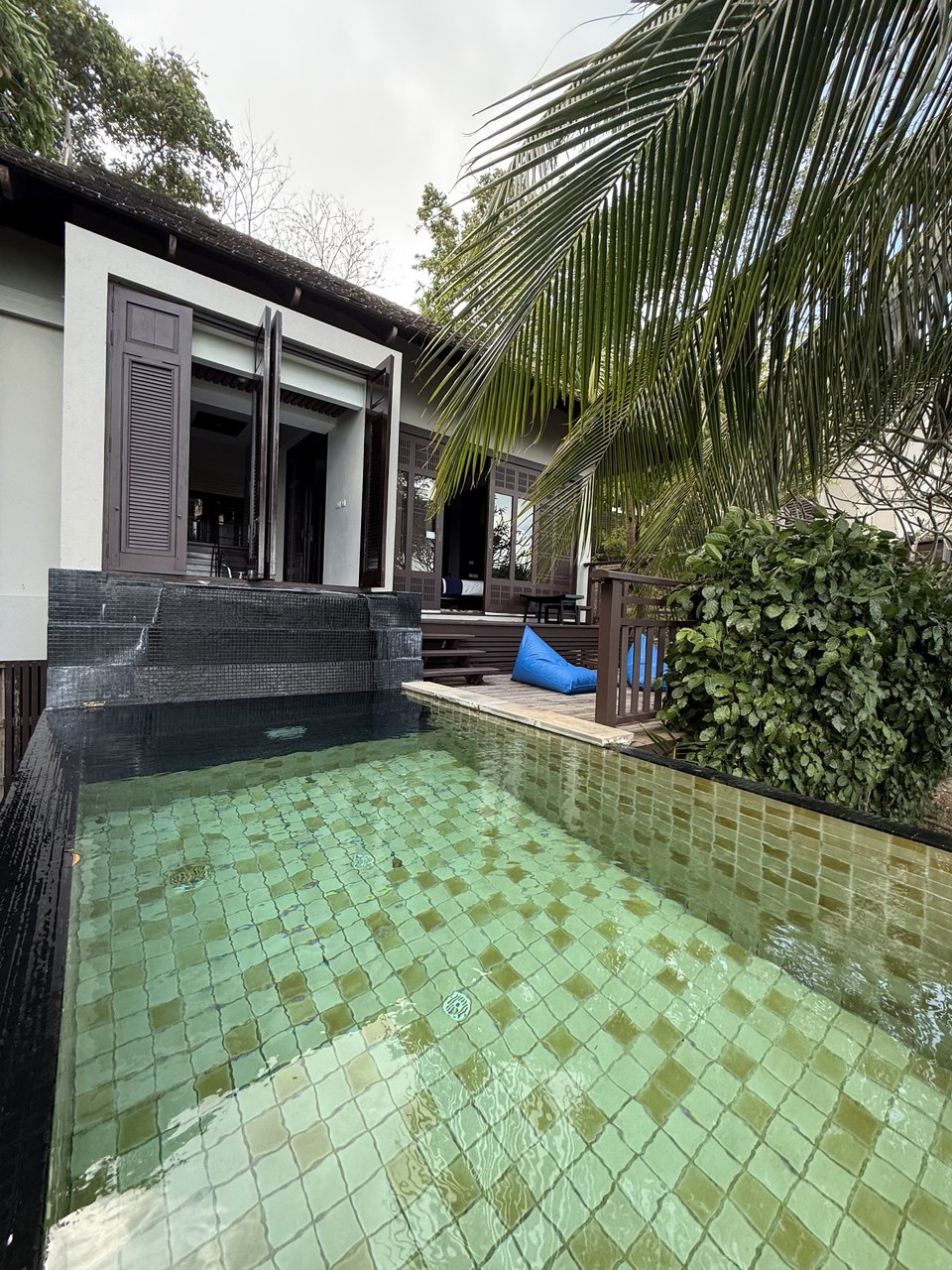 A serene pool is shown, featuring green-tinted water with tiled surfaces. Open shutters suggest a connection to the villa's interior. Lush greenery surrounds the pool area, enhancing the natural ambiance. A wooden deck is visible in the background, inviting relaxation.