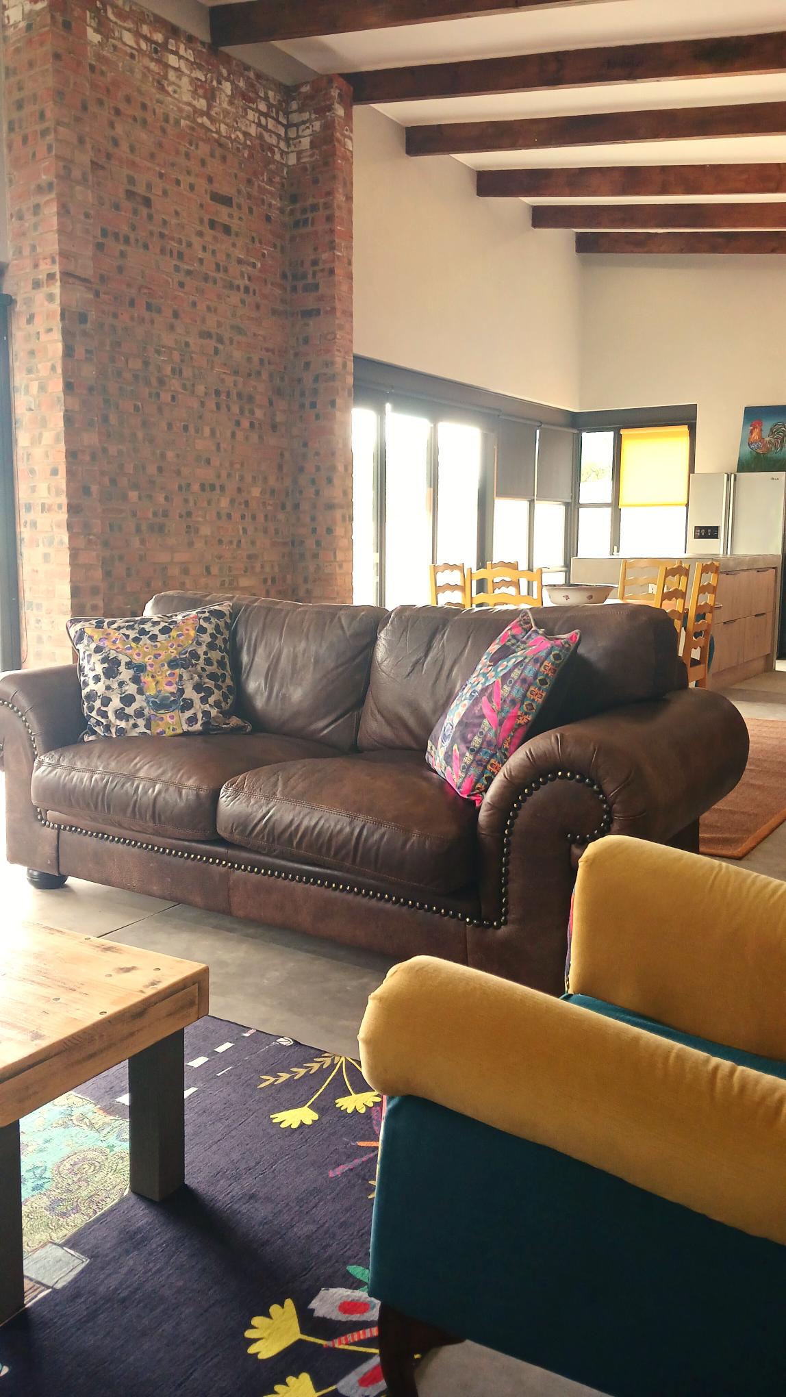 A living area showcases a leather sofa adorned with colorful cushions, complemented by a vibrant rug below. Large windows illuminate the space, providing a view towards the dining area and kitchen in the background. Natural materials, such as stone and wood, enhance the modern design.