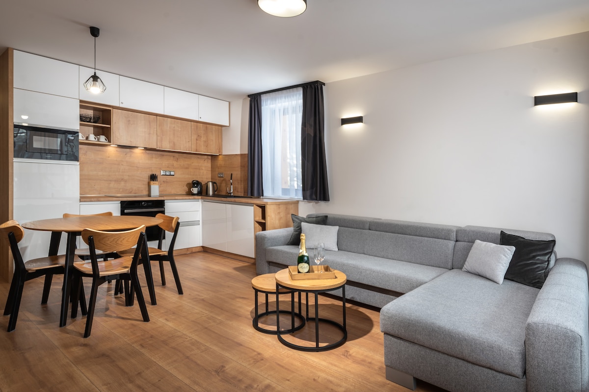 The living area features a modern design with a comfortable gray sofa and a wooden dining table accompanied by black chairs. A light wooden floor complements the stylish cabinetry in the kitchen, and a large window allows natural light to fill the space.