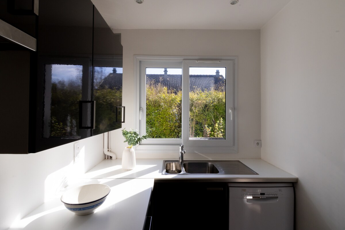 A modern kitchen is showcased with sleek black cabinetry and a white countertop. Natural light floods through a large window, illuminating the space and offering a view of greenery outside. A stainless steel sink and dishwasher are visible, adding to the kitchen's functionality.