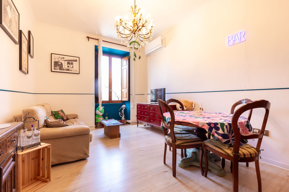 A spacious living area features a comfortable couch and a small coffee table, complemented by a dining table with colorful tablecloth and several chairs. Natural light enters through a large window, while a stylish chandelier and neon wall decor add unique touches to the vibrant space.