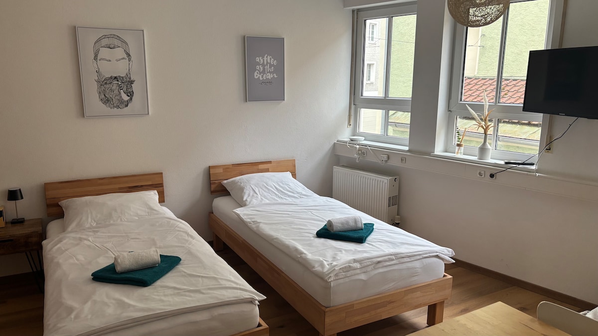 The room features two spacious beds with neatly arranged white linens and teal towels placed at the foot. Natural light fills the space through large windows, complemented by minimalistic wall decor and functional furnishings.