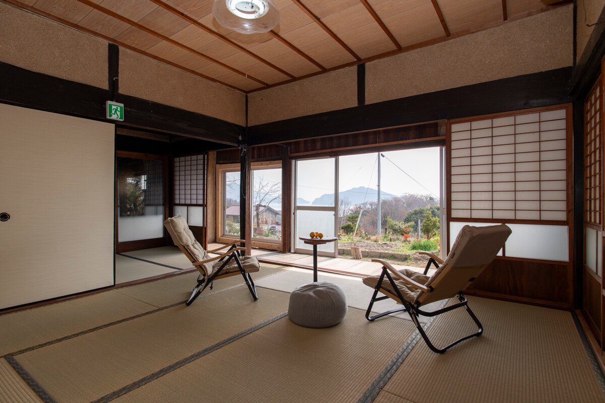 A serene space is presented with two foldable chairs positioned around a small table. Shoji screens frame the large window, which offers views of the landscape outside. Soft tatami mats cover the floor, enhancing a warm ambiance throughout the area.